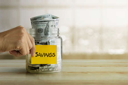 Money savings concept. Man is holding a sticky note with the word âsavingsâ stick on glass jar full of dollar banknotes.の写真素材