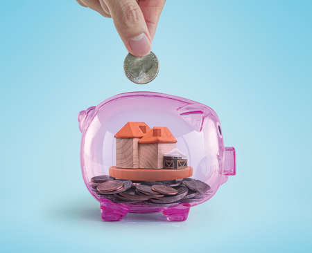 Hand putting a coin into a pink piggy bank with a house or shop toy inside.