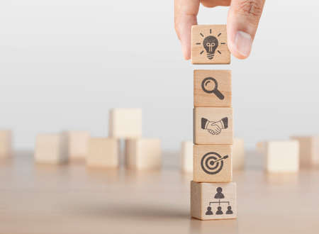 Business strategy, business management or business success concept. Hand arranging wooden blocks with business icon in tower stacked shape.の写真素材