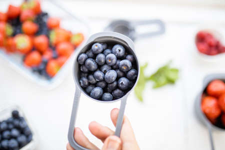 Female hand holding measuring cup full of blueberries above more berries