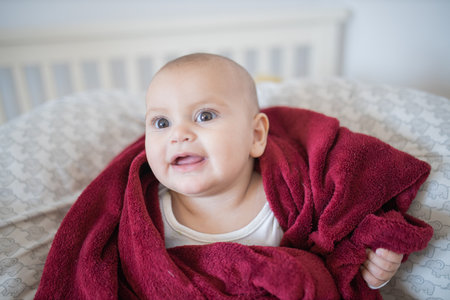 Happy baby covered with a red blanket and lying on a bed
