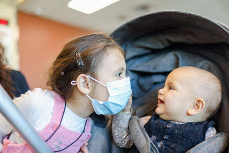 Adorable little girl wearing a face mask next to her baby sister in stroller