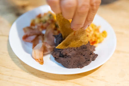 Hand picking tortilla chip from a Mexican-style eggs, bacon, and beans dishの写真素材