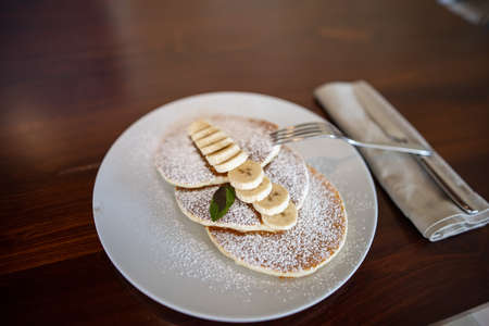 Tasty pancakes with powdered sugar and sliced banana above borwn tableの写真素材
