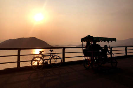 The silhouette of retro bicycle, family bike, fence, ocean, mountain and sun at sunsetの写真素材