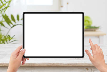 Isolated human left hand holding black tablet media device with white empty screen mockup and wooden table in kitchenの写真素材