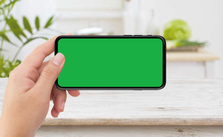 Isolated human left hand holding black mobile smart phone device mockup with green screen at indoor...