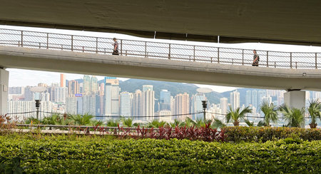 Two people walk across an elevated walkway, framed by a modern city skyline and vibrant tropical...