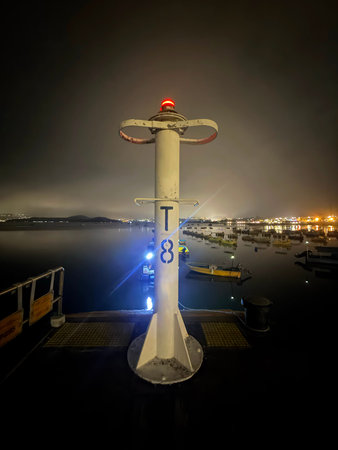 A solitary navigational post with a red light stands guard over a tranquil harbor filled with small...