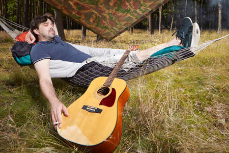 Man in wild forest relaxing in hammock bed with guitar