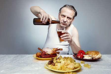 Aging man in a drinking beer and eating a lot of junk food.の写真素材