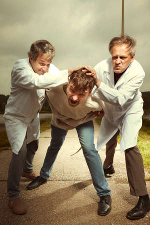 Two older retro paramedic freak hunters in medical coats catching crazy man to straitjacket