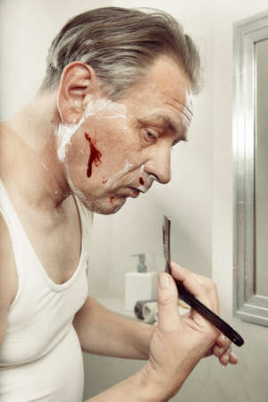 Aging man in a-shirt shaving face and cut himself with old style razorの写真素材