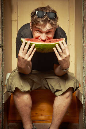 Man sitting on wooden toilet latrine and eating watermelonの写真素材
