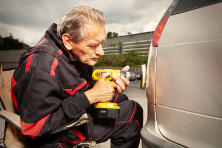 Man in overall working with drill on damage of his car directly on street