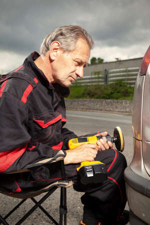 Man in overall working with drill on damage of his car directly on street