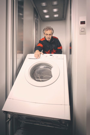 Older man in overalls moving washing machine on cart with elevatorの写真素材