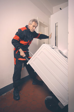 Older man in overalls moving washing machine on cart with elevatorの写真素材