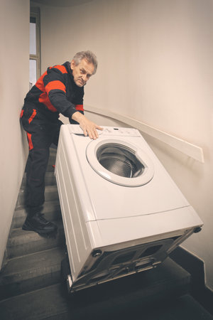 Older man in overalls moving washing machine on cart in buildingの写真素材