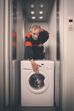 Older man in overalls moving washing machine on cart with elevatorの写真素材