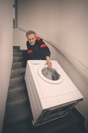 Older man in overalls moving washing machine on cart in buildingの写真素材