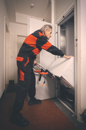 Older man in overalls moving washing machine on cart with elevatorの写真素材