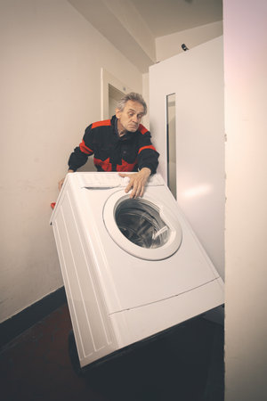 Older man in overalls moving washing machine on cart with elevatorの写真素材