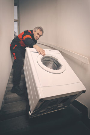 Older man in overalls moving washing machine on cart in buildingの写真素材