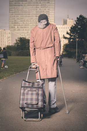 Senior citizen walking with wheel cart and stick in city parkの写真素材