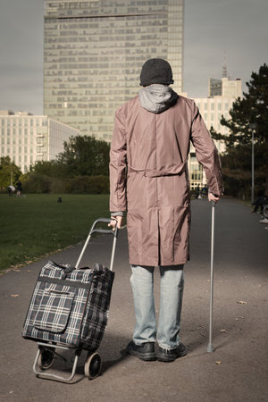Senior citizen walking with wheel cart and stick in city parkの写真素材