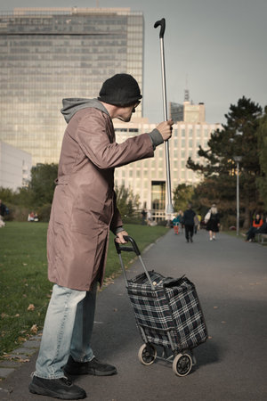 Senior citizen walking with wheel cart and stick in city parkの写真素材