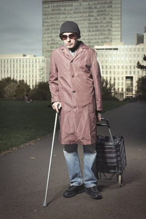 Senior citizen walking with wheel cart and stick in city parkの写真素材