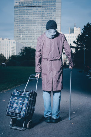 Senior citizen walking with wheel cart and stick in city parkの写真素材