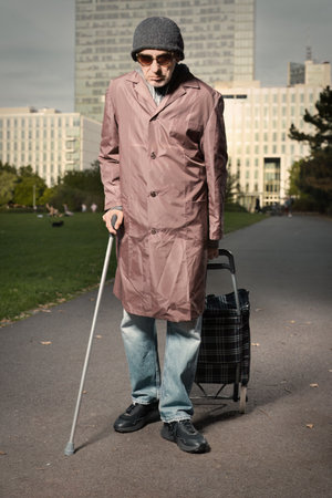 Senior citizen walking with wheel cart and stick in city parkの写真素材