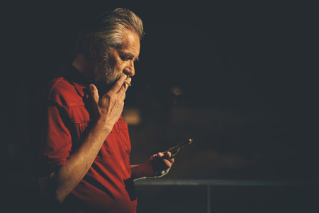 Older man in red shirt waiting for something and using mobile phone on night city locationの写真素材