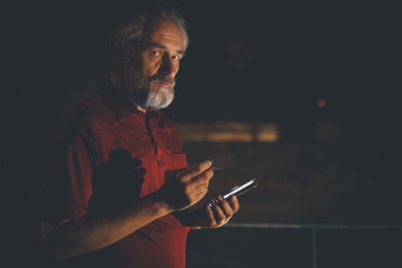 Older man in red shirt waiting for something and using mobile phone on night city locationの写真素材