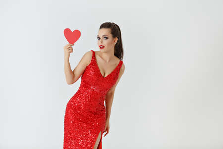 Beautiful long-haired brunette in a red dress holding a red heart. Studio, white backgroundの写真素材