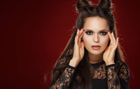 Portrait of a beautiful young brunette woman with horns on her head on a red background in studioの写真素材