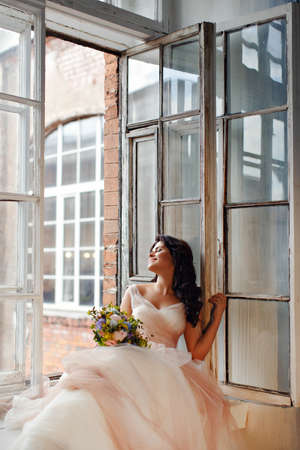 Portrait of a beautiful brunette girl in a pink dress and an air bouquet of flowers sitting on window and smiling dreamilyの写真素材