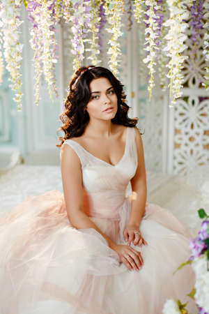 Portrait of a beautiful brunette girl in a pink dress with an air bouquet of wildflowers on a background of light and interior flowersの写真素材