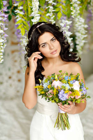 Portrait of a beautiful brunette girl in a white dress with a bouquet of wildflowers on a background of bright interiorsの写真素材