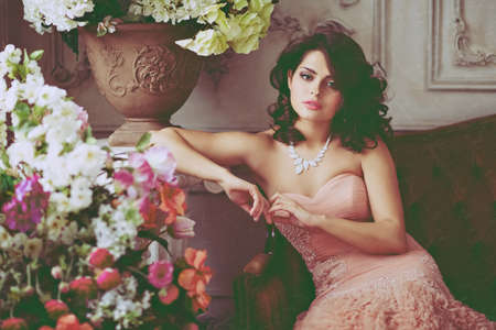 Very beautiful girl brunette in pink dress sitting on the couch surrounded by flowersの写真素材