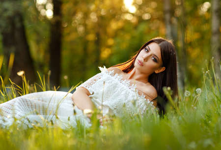 Portrait of beautiful sensual brunette girl in white dress sitting in the grass and dandelions on a summer sunsetの写真素材