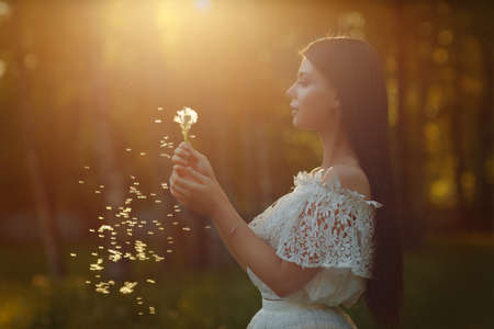 Portrait of beautiful sensual brunette girl in white dress blows on dandelion on yellow sunsetの写真素材