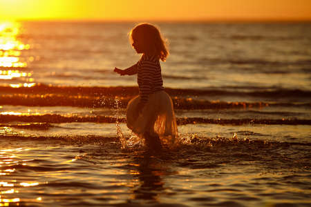 Little girl little girl in a skirt and striped sweater runs through the water, making a splash at sunset in summerの写真素材