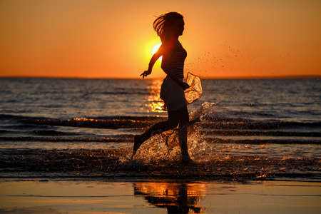 Beautiful long-haired skinny girl in a long skirt running on the water, making a splash at sunset in summerの写真素材