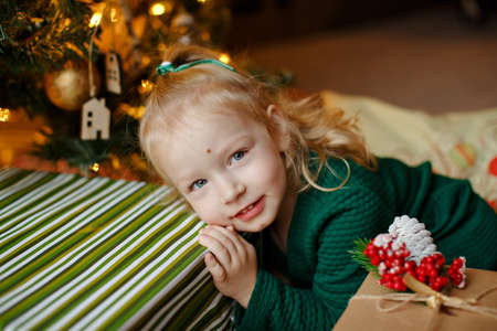 Charming chubby little blonde girl little girl in a green dress with Christmas decorations next to the giftsの写真素材