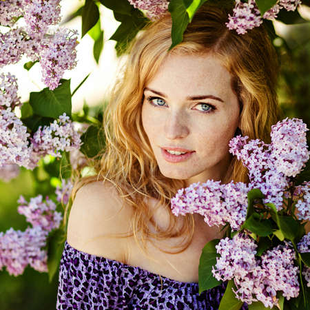 Portrait of beautiful delicate red-haired girl with freckles and clear blue eyes in the sprigs of lilac in the summer, close-upの写真素材