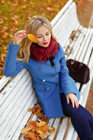 Luxury blonde girl with beautiful hair in a coat sitting on a bench in autumn parkの写真素材