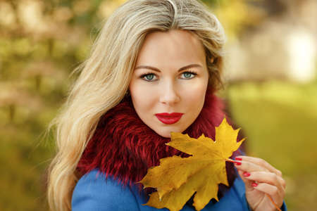 Luxury blonde girl with beautiful hair in a coat holding a maple leaf in autumnの写真素材